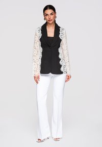 Femme portant un blazer noir avec des manches et des bords en dentelle blanche, un pantalon évasé blanc et des talons ouverts blancs, debout devant un fond uni.