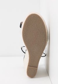 Sandale à talon compensé avec une semelle beige texturée, des brides transparentes et une finition douce. Le design comporte une bride d'accent noir pour un détail supplémentaire.