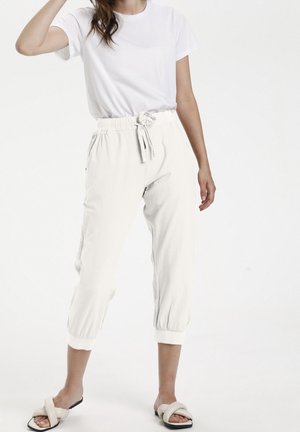 Kvinna i vit kortärmad t-shirt, vita knytbyxor i cropped-stil och vita platta sandaler på en enfärgad bakgrund.