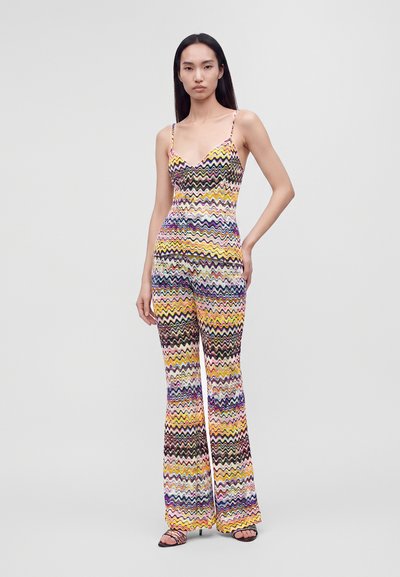 Femme aux longs cheveux foncés portant un haut sans manches à motif zigzag coloré et un pantalon évasé assorti, debout avec une main sur la hanche.