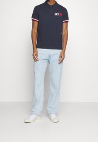 Marineblauwe polo met contrasterende rode en witte gestreepte mouwen, voorzien van een grafische opdruk "1985 NEW YORK" op de borst. Licht gewassen jeans en witte sneakers.