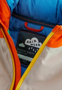 Interior multicolor de una chaqueta de lluvia con forro azul, detalles en naranja y amarillo, y una etiqueta con un diseño de nube y lluvia.