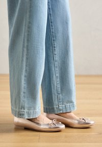 Metalliska roséguld ballerinas med rund tå, liten rosettdetalj, kombinerade med ljusblå vida jeans, på ett trägolv.