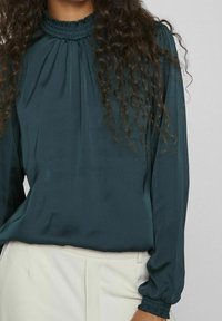 Ciemnozielona bluzka z wysokim marszczonym kołnierzem, długimi rękawami zakończonymi elastycznymi mankietami oraz miękką, gładką teksturą. W zestawie z jasnymi spodniami.