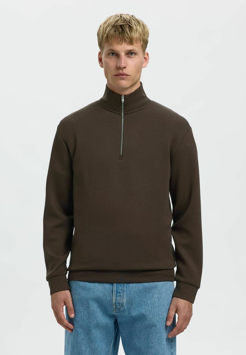 Hnědá mikina na zip s vysokým límcem, vyrobená z hladké tkaniny. Má dlouhé rukávy a volný střih. Nosená s světle modrými džíny.