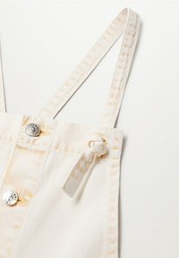 Les salopettes en denim blanc sont dotées de bretelles ajustables, d'un détail noué et de ferrures argentées. Des coutures subtiles ajoutent de la texture au tissu.