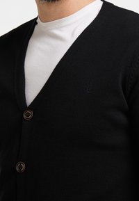 Cardigan nero a maglia con scollo a V, dotato di un piccolo logo ad ancora, due bottoni marroni e una texture liscia sopra una camicia bianca sottostante.