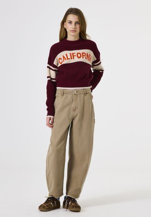 Bordeaux gebreide trui met de tekst "CALIFORNIA", crèmekleurige strepen; beige wijde broek; bruine schoenen met een luipaardprint; ontspannen pasvorm.
