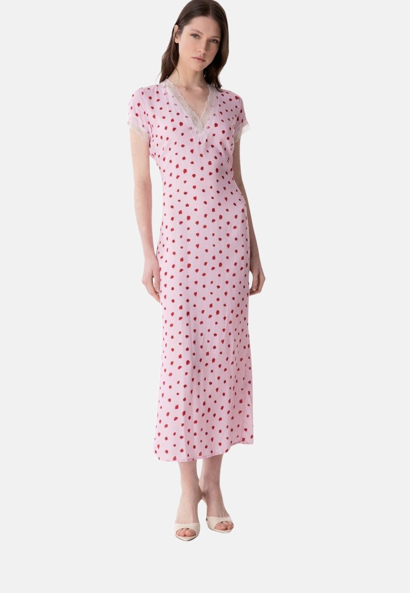 Frau trägt ein pastellrosa Maxikleid mit roten Punkten, Spitzen-V-Ausschnitt, kurzen Ärmeln und beigefarbenen offenen High Heels, steht vor weißem Hintergrund.