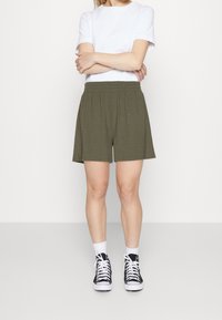 Shorts côtelés vert olive avec une taille élastique, associés à un t-shirt blanc et des baskets noires, se tenant sur un fond uni.