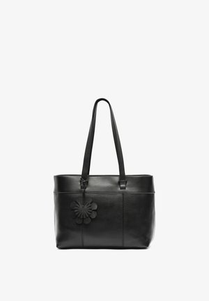 Sort læder tote bag med lange stropper og en sort blomstformet charme hængende nær det øverste venstre hjørne.