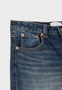 Zbliżenie na ciemnoniebieskie dżinsy z denimu, pokazujące przednią kieszeń z miedzianymi nitami, szlufką na pasek i kontrastującym przeszyciem na jasnoszarym tle.