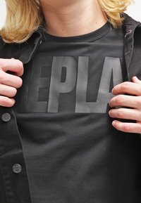 Black T-shirt with large matte black "REPLAY" text on the front, made of cotton, featuring a round neck and short sleeves.