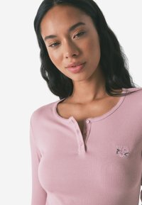 Top lungo a maniche lunghe di un leggero rosa con colletto a scollo a V, cinque bottoni e dettagli floreali ricamati sul lato sinistro del petto. Tessuto morbido.