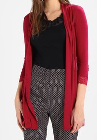 Vínový cardigan s otevřeným předním dílem, tříčtvrteční krajkové rukávy, v kombinaci s černým topem s krajkovým lemem a vzorovanými kalhotami s malými motivy.