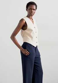 Beige vest with a deep V-neck and five buttons, paired with dark navy wide-leg trousers. Smooth fabric, tailored fit, minimalistic design.