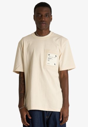 Giovane uomo con capelli corti indossa una T-shirt oversize beige con una tasca bianca stampata e jeans blu scuro, in piedi davanti a uno sfondo bianco.