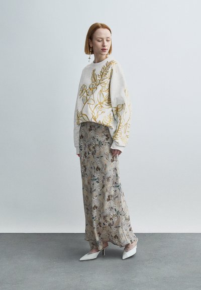 Γυναίκα με λευκό πουλόβερ με χρυσή κεντημένη φυλλωσιά, μακριά φλοράλ φούστα και λευκές μυτερές γόβες, στέκεται σε γκρι πάτωμα μπροστά σε παστέλ τοίχο.