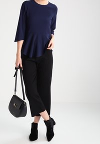 Top peplo blu navy a tre quarti, abbinato a pantaloni neri a vita alta e stivali neri alla caviglia, mentre tiene una borsa nera.