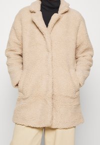 Beige, pluizige jas met een zachte, gestructureerde oppervlakte, een reverskraag en twee voorzakken. Het ontwerp zonder knopen zorgt voor een eenvoudig silhouet.