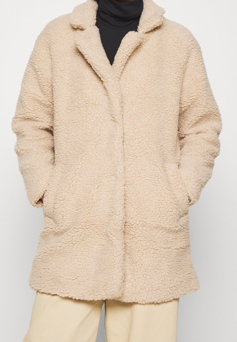 Beige, pluizige jas met een zachte, gestructureerde oppervlakte, een reverskraag en twee voorzakken. Het ontwerp zonder knopen zorgt voor een eenvoudig silhouet.