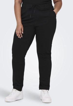 Persoon draagt zwarte nauwsluitende broek met trekkoord in de taille en witte sneakers, staat met één hand in de zak en de andere hand langs het lichaam.