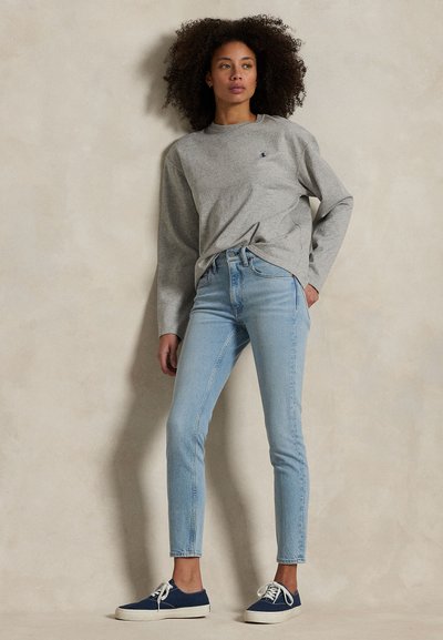 Moteris su garbanotais plaukais, vilkinti pilką ilgomis rankovėmis marškinėlį, šviesiai mėlynas siauras džinsus ir tamsiai mėlynas sportbačius, stovinti prie paprastos smėlio spalvos sienos.