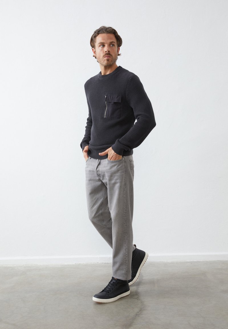 Pull à côtes sombre avec une poche zippée, pantalon gris clair et baskets montantes noires, debout sur un sol en béton devant un fond sobre.