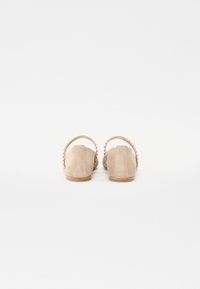 Chaussures slip-on en daim beige avec un petit talon et un dos arrondi, comportant une sangle festonnée le long du bord pour un détail supplémentaire.