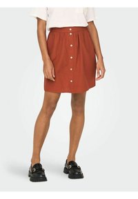 Gonna di cotone color ruggine con vita arricciata, caratterizzata da bottoni frontali e taglio al ginocchio. Indossata con mocassini neri e una maglietta bianca.