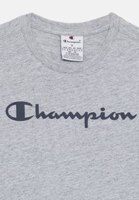T-shirt grigio in cotone con scollatura a girocollo, caratterizzato da un grande logo blu scuro "Champion" sul petto. Contiene un'etichetta di cura.