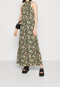 Maxikleid aus leichtem Stoff mit einem Halsband, das ein grünes und braunes Blättermuster auf einem cremigen Hintergrund zeigt. Schwarze Plateau-Sandalen.