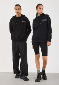 Zwei Personen mit geflochtenen Haaren, die schwarze Hoodies mit weißem Schriftzug tragen, kombiniert mit lockeren Hosen und Radlerhosen, schwarzen Stiefeln und gestreiften Socken.