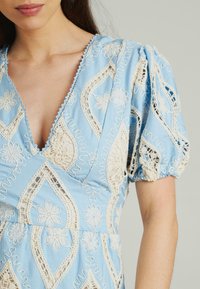 Hellblaues Kleid mit filigraner weißer Stickerei in Blumen- und Paisleymustern. Puffärmel und gewellte Saumdetails heben das Design hervor.