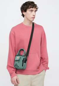 Ροζ φούτερ με μπροστινή τσέπη, φορεμένο με μια μικρή πράσινη τσάντα που διαθέτει πάνω λαβή, ρυθμιζόμενο λουρί και φερμουάρ λεπτομέρειες.