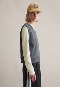 Grauer ärmelloser Pullover über einem weißen langärmligen Hemd, kombiniert mit schwarzen Hosen mit weißen Streifen. Hellbraune Mütze mit der Nummer 15.