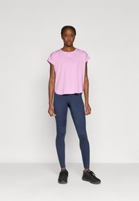 Ροζ αθλητική μπλούζα με κοντά μανίκια, στρογγυλεμένο τελείωμα και λογότυπο, συνδυασμένη με μπλε σκούρο κολάν και μαύρα αθλητικά παπούτσια· απαλή υφή, εφαρμοστή σχεδίαση.