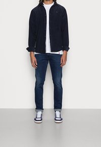 Camisa de corduroy azul marino de botón sobre una camiseta blanca, combinada con unos pantalones vaqueros azul oscuro y zapatillas blancas con detalles en azul marino.
