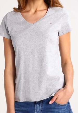 Femme portant un T-shirt gris clair à col en V et manches courtes avec un petit logo sur la poitrine, associé à un jean bleu et une main dans la poche.