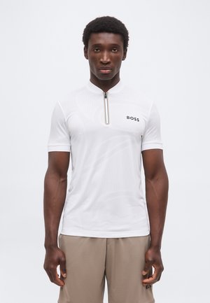 Άνδρας που φοράει λευκό κοντομάνικο πουκάμισο με φερμουάρ και λογότυπο "BOSS" και μπεζ παντελόνι, στέκεται μπροστά σε ένα απλό φωτεινό φόντο.