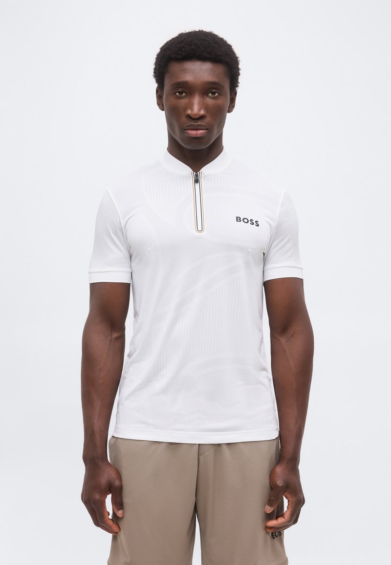 Άνδρας που φοράει λευκό κοντομάνικο πουκάμισο με φερμουάρ και λογότυπο "BOSS" και μπεζ παντελόνι, στέκεται μπροστά σε ένα απλό φωτεινό φόντο.