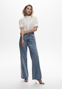 Blusa blanca bordada con detalles de volantes, combinada con vaqueros de pierna ancha de cintura alta y sandalias de tacón transparente.