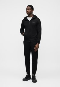 Melns zip-up džemperis ar kapuci un atbilstošas bikses no mīksta auduma. Džemperim uz krūtīm ir uzšūta logo. Modelis ir ģērbies melnās sporta kurpēs.