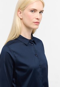 Camicia azzurro navy a maniche lunghe con bottoni, realizzata in tessuto morbido, dotata di colletto tradizionale e chiusura frontale con bottoni. Presenta una leggera lucentezza.