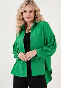 Grüne Button-Down-Bluse aus glattem Stoff mit lockerer Passform, abgesenkten Schultern und gerafften Bündchen. Kombiniert mit einem schwarzen Oberteil und einer Perlenkette.