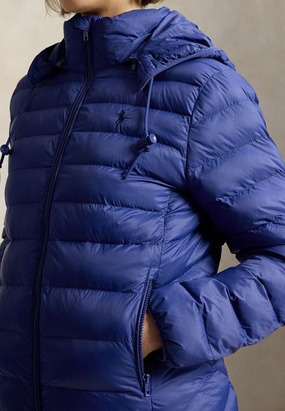 Γυναίκα που φοράει μπλε καπιτονέ μπουφάν με κουκούλα και κορδόνια, με τα χέρια στις πλαϊνές τσέπες με φερμουάρ, στραμμένη ελαφρώς αριστερά.