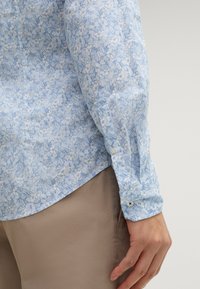 Camicia floreale azzurro chiaro con polsini abbottonati, caratterizzata da un motivo delicato e una texture liscia. Abbinata a pantaloni beige.