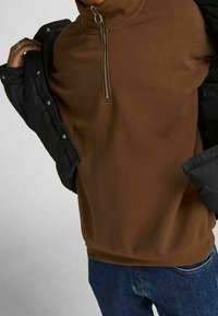 Sudadera de felpa marrón con cuello alto y detalle de cremallera plateada. Combinada con una chaqueta negra. Los jeans son azules y de corte relajado.