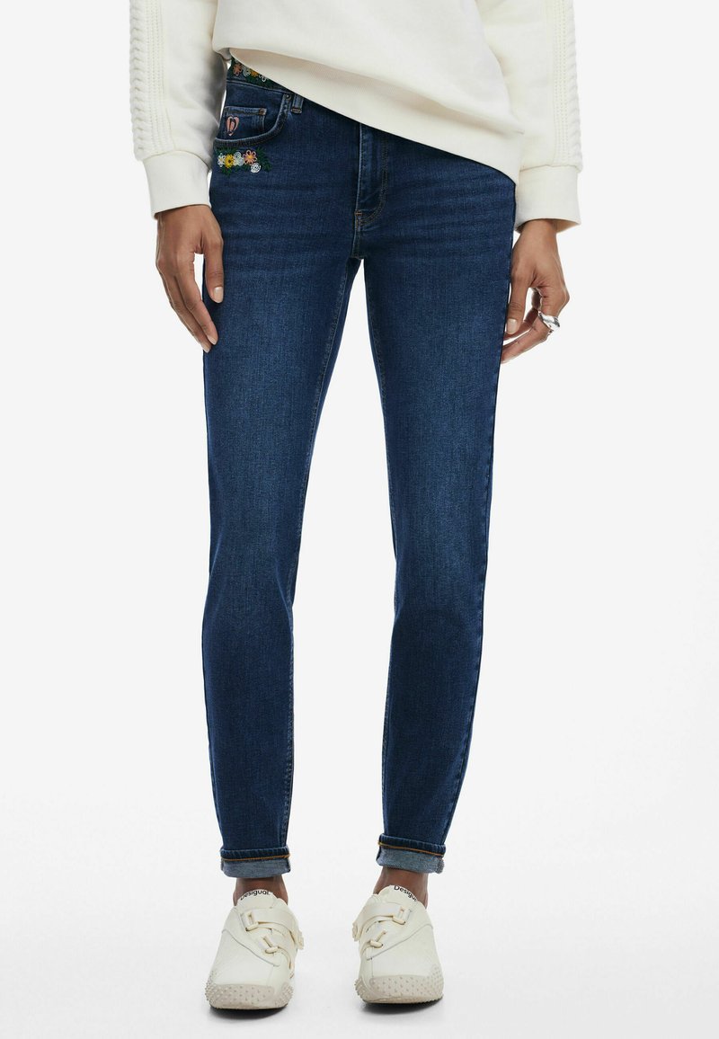 Jeans skinny in denim blu scuro con ricami floreali sulla tasca anteriore; abbinati a un maglione chiaro e sneakers bianche con cinturini.