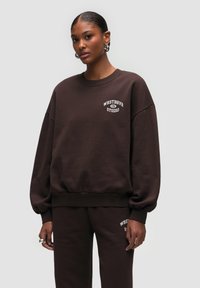 Felpa marrone con colletto a girocollo e vestibilità rilassata, caratterizzata da un logo ricamato bianco "WESTBHVR STUDIO" sul petto. Tessuto morbido.